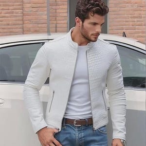 Peut inclure: Veste en cuir blanche avec un motif texturé, fermeture éclair et poches latérales. La veste est portée avec un t-shirt blanc, un jean bleu et une ceinture marron. Une montre est visible au poignet.
