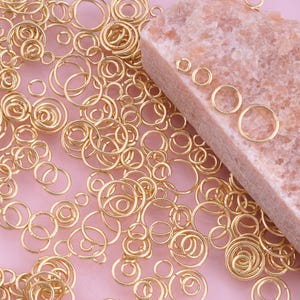 May include: A collection of gold-colored jump rings and spiral rings, scattered on a pink surface. Several rings are arranged on a rectangular, light-brown stone. The rings vary in size and shape, suitable for jewelry making and crafts.