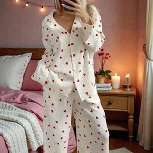 May include: Cream-colored pajama set with a repeating pattern of small red hearts. The set includes a button-down shirt with a collar and long pants. The pajamas are made of a soft, lightweight fabric, perfect for lounging or sleeping.