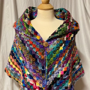May include: A colorful crocheted shawl with a large collar and a wooden button closure. The shawl is made with a variety of different colored yarns, creating a patchwork effect.