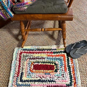 Puede incluir: Una alfombra rectangular hecha a mano, con un diseño geométrico colorido. La alfombra presenta un rectángulo central granate rodeado de bordes concéntricos de varios colores. Una silla de madera con un cojín verde y una manta colorida están en el fondo.