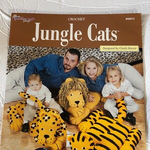 May include: A crochet pattern book titled "Jungle Cats" with a family and three crocheted animal toys: a lion, a tiger, and a leopard. The cover text reads "Friendly Felines Make Perfect Playtime Companions, Each Made Using Worsted Yarn."