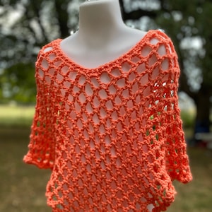 May include: A coral orange crochet poncho with a delicate openwork pattern. The poncho is displayed on a white mannequin against a blurred green background.