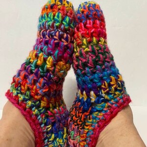 May include: A pair of colourful crocheted slipper socks with a pink trim. The socks are made with a variety of different coloured yarns, creating a rainbow effect.