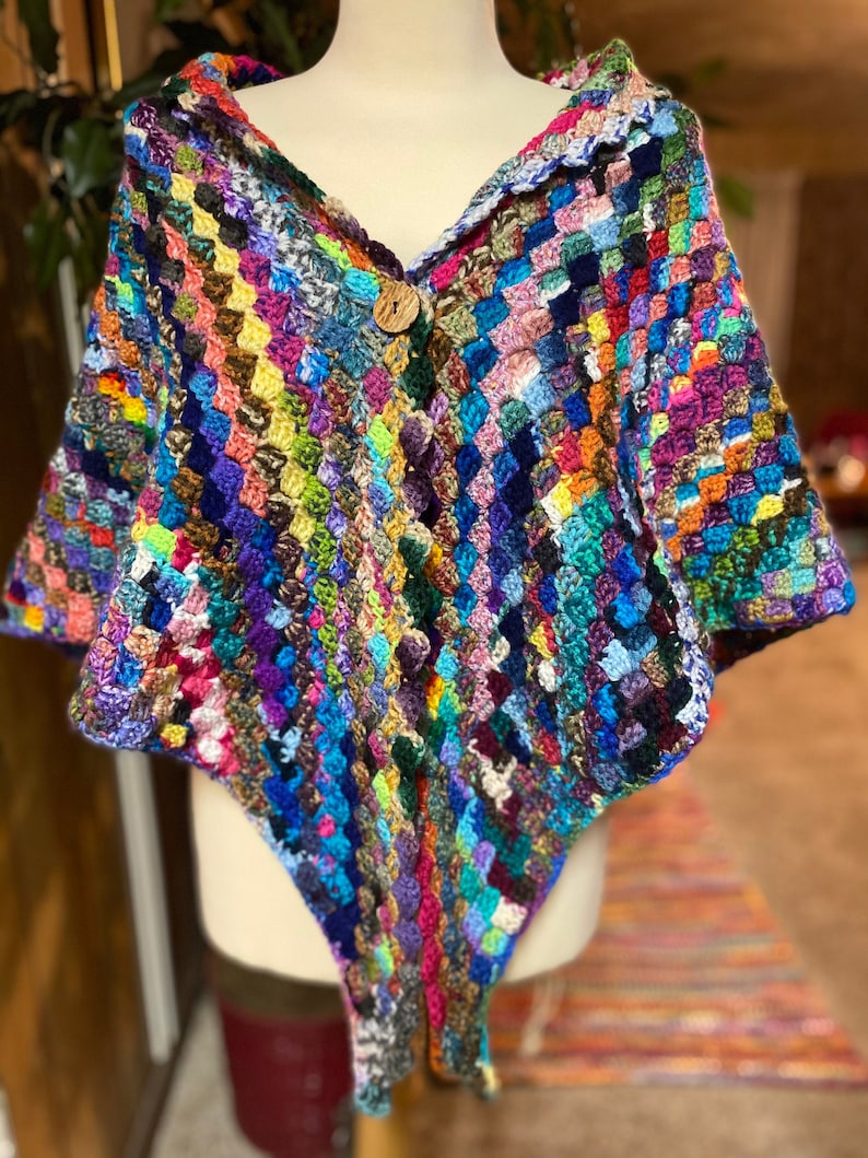 May include: A colorful crocheted shawl with a wooden button closure. The shawl is made with a variety of different colored yarns and has a unique, textured design.