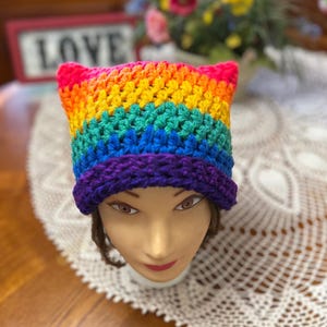 May include: A handmade, rainbow-colored crochet hat with cat ears. The hat features bands of red, orange, yellow, green, blue, and purple. The hat is displayed on a mannequin head, with a blurred background of flowers and a sign that says "LOVE."