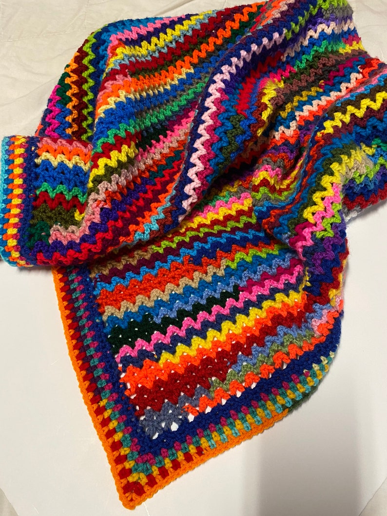 May include: A colorful crocheted blanket with a zig-zag pattern and a border of alternating colors. The blanket is made with a variety of bright colors, including red, orange, yellow, green, blue, purple, and pink.