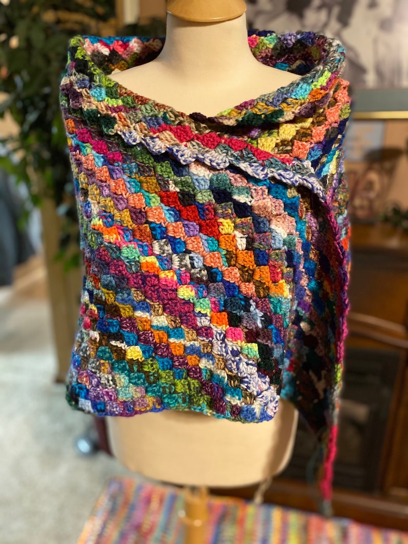 May include: A colorful crocheted shawl with a variety of colors, including blue, green, red, and purple. The shawl is draped over a mannequin and has a fringed edge.
