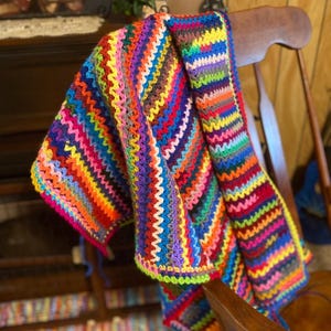 May include: A colorful crocheted blanket draped over a wooden rocking chair. The blanket is made with a variety of bright colors, including red, orange, yellow, green, blue, purple, and pink.