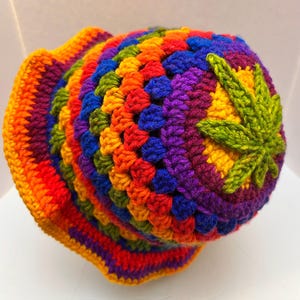 May include: A crochet bucket hat featuring a vibrant rainbow color palette of red, orange, yellow, green, blue, and purple yarn. The hat is constructed with a textured granny square stitch pattern and includes a prominent green leaf applique.