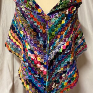 May include: A colorful crocheted shawl with a large collar and a wooden button closure. The shawl is made with a variety of different colored yarns, creating a patchwork effect.