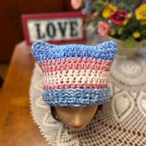 May include: A handmade crochet hat in blue, pink, and white, with ear-like points at the top. The hat is displayed on a mannequin head. The word "LOVE" is visible in the background.