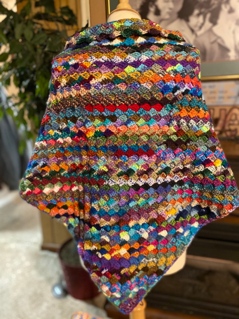 May include: A colorful crocheted shawl with a variety of colors, including blue, green, purple, and red. The shawl is made with a granny square stitch and has a triangular shape.