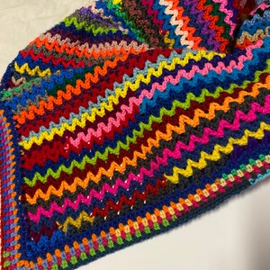 May include: A colorful crocheted blanket with a zig-zag pattern in shades of red, orange, yellow, green, blue, purple, pink, and brown. The blanket has a dark blue border with a multi-colored trim.