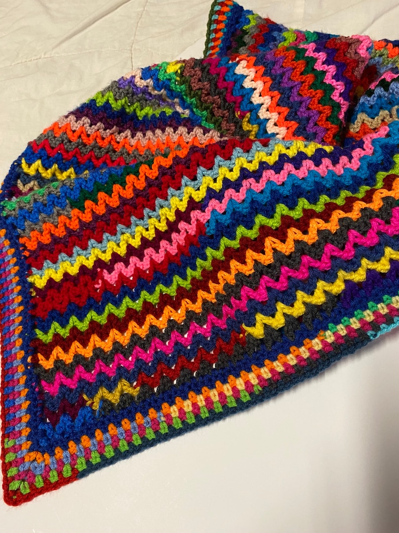 May include: A colorful crocheted blanket with a zig-zag pattern in shades of red, orange, yellow, green, blue, purple, pink, and brown. The blanket has a dark blue border with a multi-colored trim.