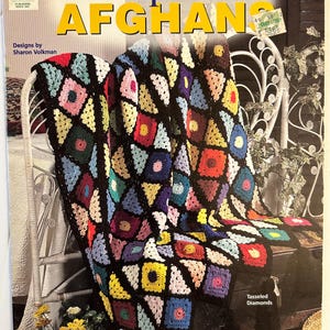 May include: A craft book titled Crochet Scrap Yarn AFGHANS featuring a multi-coloured afghan made of crocheted squares in shades of red, yellow, blue, pink, and green, joined by black yarn. The cover displays the text Designs by Sharon Volkman.