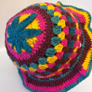May include: A crochet bucket hat featuring a vibrant granny square pattern in shades of deep burgundy, bright yellow, hot pink, and teal. The crown is topped with a prominent, star-shaped teal crochet applique, and the brim has a wavy edge.