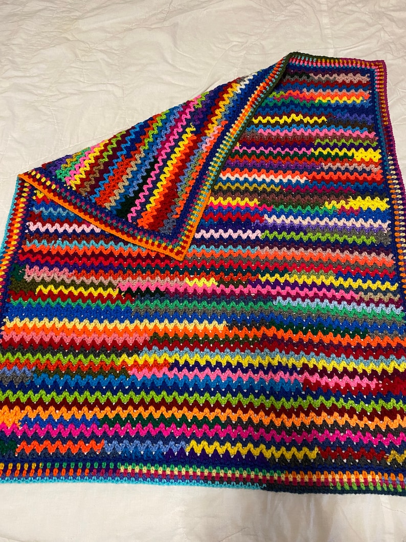 May include: A colorful crocheted blanket with a wavy pattern. The blanket is made with a variety of bright colors, including red, orange, yellow, green, blue, purple, and pink.