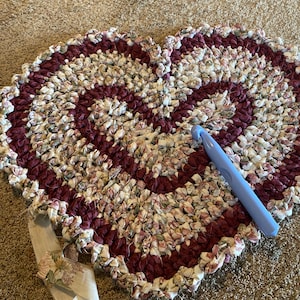 May include: A heart-shaped crocheted rug made from fabric scraps in shades of burgundy, white, and pink. A blue crochet hook is lying on the rug.