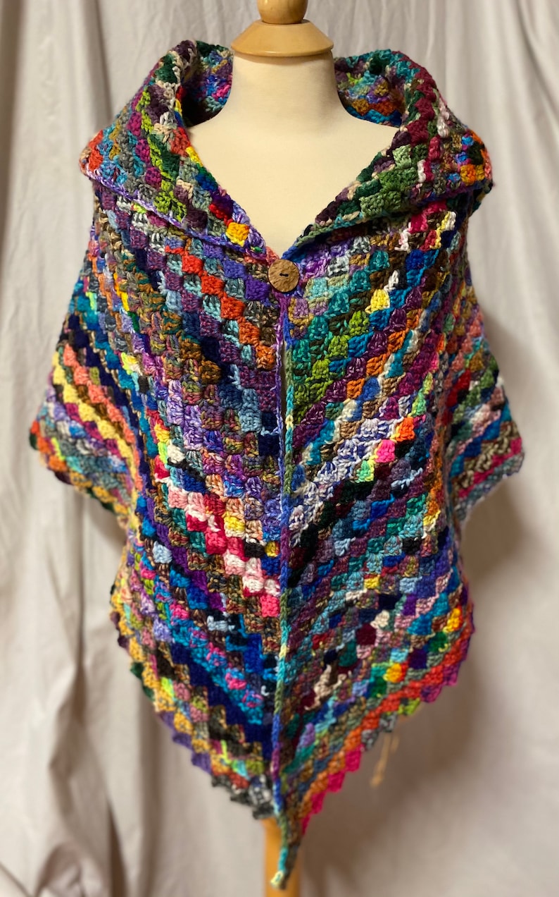 May include: A colorful crocheted shawl with a large collar and a wooden button closure. The shawl is made with a variety of different colored yarns, creating a patchwork effect.