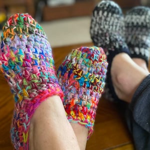 May include: A pair of colourful crocheted slippers with a multi-coloured yarn design. The slippers are worn on a person's feet.