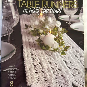 May include: A crochet pattern book featuring 8 table runner designs. The cover shows a white crocheted table runner with a candle and flowers on a table. The book title is "Table Runners in half the time!" and the text "Made with sizes 3 and 5 cotton thread." is also visible.