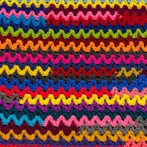 May include: A colorful crocheted blanket with a wavy pattern. The blanket is made with a variety of bright colors, including red, orange, yellow, green, blue, purple, and pink.
