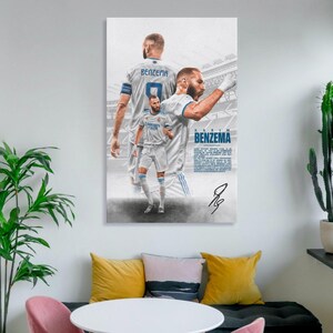 Może przedstawiać: Oprawiony plakat z Karimem Benzemą w białym stroju piłkarskim. Plakat zawiera wiele zdjęć zawodnika, nazwisko "BENZEMA" i jego podpis. Tło przedstawia stadion.