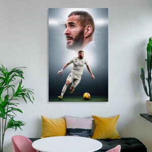 以下が含まれることがあります： 白いユニフォームを着たサッカー選手が、緑のフィールドで黄色のサッカーボールを蹴っている様子を描いた額入りのプリント。画像には、アクションショットの上に選手の肖像画が含まれています。背景はスタジアムです。