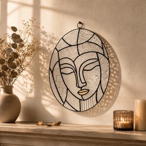 Könnte beinhalten: Ein ovales Wandbild aus Buntglas, das ein stilisiertes menschliches Gesicht mit geschlossenen Augen und einem goldfarbenen Mund zeigt. Das Stück besteht aus strukturierten, lichtdurchlässigen Glasscheiben, die durch schwarze Metalllinien verbunden sind.