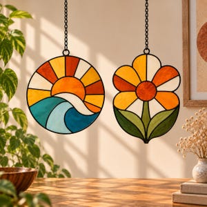 Könnte beinhalten: Zwei Buntglas-Sonnenfänger, die an schwarzen Ketten hängen. Eines ist ein kreisförmiges Stück, das eine Sonne über blauen Meereswellen in Orange, Gelb und Türkis darstellt. Das andere ist ein blumenförmiges Stück mit orangen Blütenblättern und grünen Blättern.