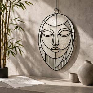 May include: An oval stained glass wall hanging featuring a stylized human face composed of clear, textured glass panels outlined in black metal. The design includes geometric shapes and lines forming closed eyes, a nose, and lips, suspended by a ring.