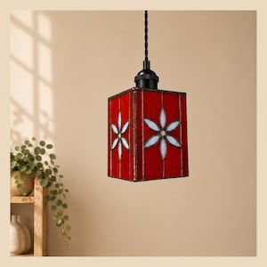 Könnte beinhalten: Eine quadratische Hängeleuchte mit roten Buntglas-Paneelen, die auf jeder Seite ein weißes, sechsblättriges Blumenmuster aufweisen. Die Lampe ist an einem schwarzen Kabel aufgehängt und besitzt eine schwarze Metallfassung oben am Glasschirm.
