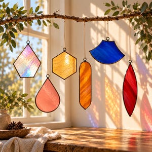 May include: Six geometric stained glass suncatchers hang from a branch. The shapes include an iridescent pentagon, a pink teardrop, a yellow hexagon, an orange rectangle, a blue fan, and a red leaf shape, all framed in thin black metal.