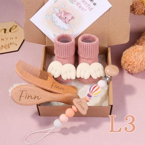 Puede incluir: Un juego de regalo para bebés en una caja de cartón. El juego incluye calcetines rosas para bebés con detalles de alas blancas, un peine y un cepillo de madera, y un clip para chupete con un diseño de globo aerostático. El peine y el cepillo están grabados con el nombre "Finn".