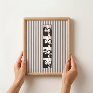 May include: A wooden framed photo strip display with a grey and white striped background. The photo strip contains five black and white images of two people. The frame is held by two hands with pink nail polish.