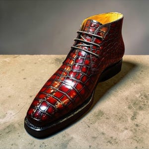 Könnte beinhalten: Ein einzelner, hochwertiger Stiefel aus rotem und braunem Leder mit einem strukturierten, krokodilartigen Muster. Der Stiefel hat eine runde Spitze, einen Schnürverschluss und eine schwarze Sohle. Die Innenseite ist mit gelbem Leder gefüttert.