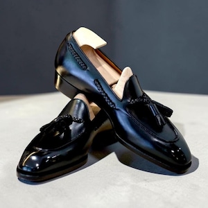 May include: A pair of black leather loafers featuring a polished finish, a braided leather trim along the collar, and a decorative tassel on the vamp. Each shoe includes a light-colored wooden shoe tree inserted inside to maintain its shape.