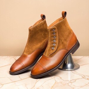 May include: A pair of brown leather ankle boots. The boots feature a combination of smooth and suede leather, with button detailing on the side. The toe caps are rounded, and the boots are set on a marble surface.