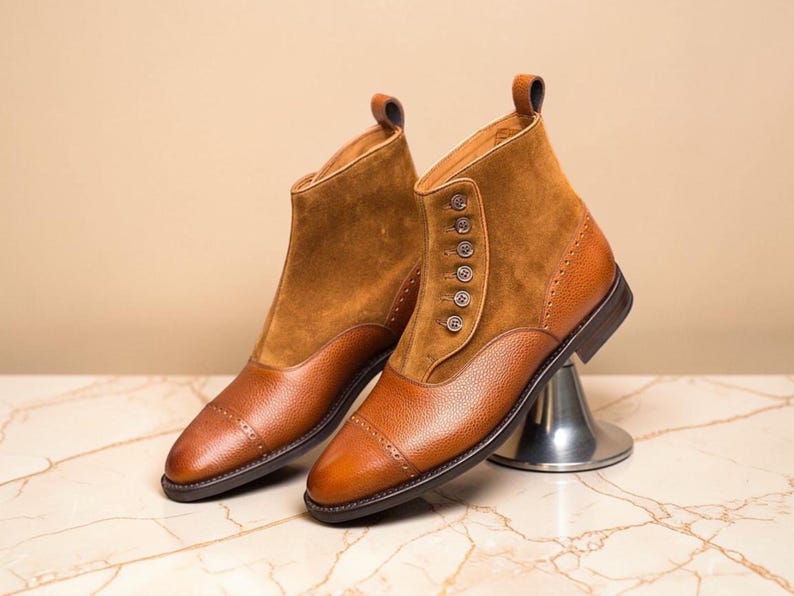 May include: A pair of brown leather ankle boots. The boots feature a combination of smooth and suede leather, with button detailing on the side. The toe caps are rounded, and the boots are set on a marble surface.