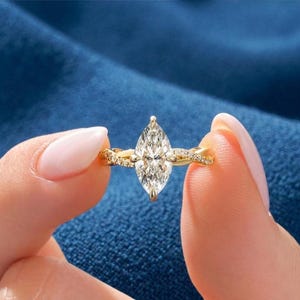 May include: A gold engagement ring featuring a marquise-cut diamond. The ring has a twisted band with small accent stones. The diamond is held in place by four prongs. The ring is held against a blue backdrop.