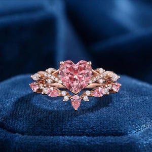 Op de afbeelding: Een roségouden ringenset met een hartvormige roze edelsteen als middelpunt. De ring is versierd met kleinere roze en heldere edelstenen, wat een sprankelend en elegant ontwerp creëert. De ring ligt op een donkerblauwe fluwelen ondergrond.