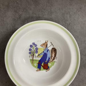 May include: White ceramic bowl with a green rim and a whimsical illustration of a squirrel in blue overalls carrying a rake and watering can. The squirrel is in a garden scene with flowers and a fence.