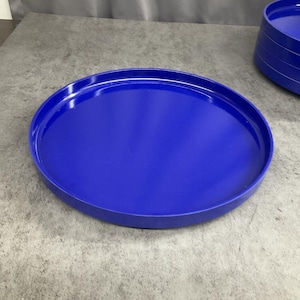 May include: A round, vibrant blue tray with a smooth, glossy finish and a raised edge. The tray features a simple, circular design with a flat base and a uniform, solid color throughout. A stack of identical blue trays is visible in the background.