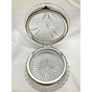 May include: A clear, round, hinged glass trinket box with a silver-colored metal rim. The lid is open, revealing etched floral designs. The base features a starburst pattern. The box is likely used for storing small jewelry or keepsakes.