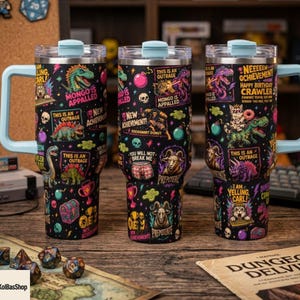 May include: Three black stainless steel tumblers with light blue lids and handles, featuring a colorful pattern of dinosaurs, skulls, trophies, and text including "Mongo is Appalled," "New Achievement," "This is an Outrage," and "You Will Not Break Me."