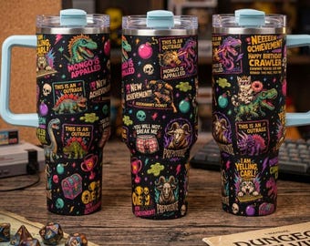 May include: Three black stainless steel tumblers with light blue lids and handles, featuring a colorful pattern of dinosaurs, skulls, trophies, and text including "Mongo is Appalled," "New Achievement," "This is an Outrage," and "You Will Not Break Me."