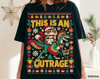 May include: A black t-shirt featuring a graphic print of a fluffy cat wearing a Santa hat and riding a colorful dinosaur. The design includes the text THIS IS AN OUTRAGE and RIDE OR ROAR, surrounded by festive patterns of snowflakes, trees, and crowns.