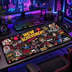 May include: A rectangular black desk mat featuring a vibrant, multi-colored graphic print with the text NEW ACHIEVEMENT! and various cartoon illustrations including a treasure chest, a dinosaur, cats, and humorous phrases like YOU WILL NOT BREAK ME.