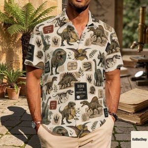 May include: A short-sleeve button-down shirt featuring an all-over print of various dinosaurs, fossils, and ferns in shades of brown, green, and cream. The shirt includes rectangular text boxes with the phrases Ancient Giants, Stay Wild, and Born to be Prehistoric.